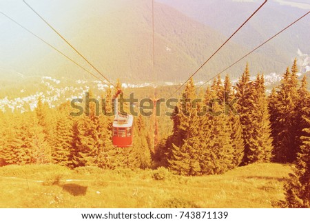 A toned picture of bright red cableway cabin moving to the top of the mountain over the beautiful landscapes with blue mountains, forest, green valley and a town at the background in warm sunlight.