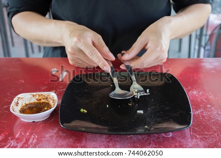 Man wearing black casual wear holding fork and spoon with empty dirty black plate and white chili sauce cup after eaten on red table background