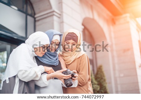 Traveling and photography. Young muslim women in beautiful hijab holding the camera and looking into it. They are smiling. Outdoors in town.