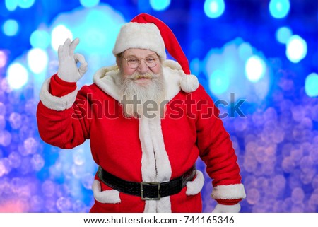 Happy Santa Claus showing ok sign. Smiling Santa Claus showing ok gesture standing on blue shiny background. Aged Santa Claus doing gesture.