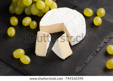 Cutting board of camembert cheese and white grapes on dark stone background.