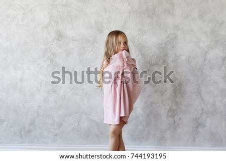 Adorable cute baby girl with long straight hair posing at blank grey wall wearing beautiful pink dress, having shy look. Sweet female kid trying on fancy dress. Kids, fashion and style concept