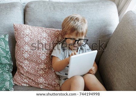 Curious funny adorable little girl wearing big spectacles having rest indoors, sitting on sofa and using touchpad pc, watching favourite cartoons online. Kids, childhood and modern technologies