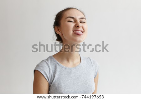Naughty girl misbehaving, sticking out her tongue at camera as a sign of disobedience, protest and disrespect. Picture of Caucasian young woman grimacing and having fun at blank studio wall