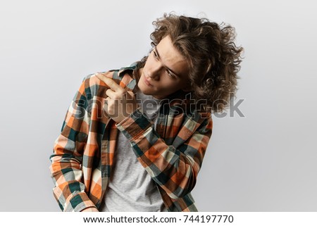 Loot at this. Isolated portrait of handsome young male customer dressed in trendy clothes looking sideways and pointing index finger, showing something interesting on blank white studio wall