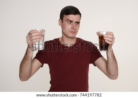 Isolated picture of indecisive young Caucasian man with dark hair frowning, having doubtful uncertain expression, facing dilemma, holding two glasses, choosing between water and alcohol drink