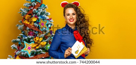 Festive season. Portrait of happy young woman near Christmas tree on yellow background with envelope and postcard