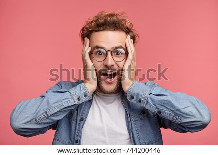Indoor shot of amazed excited man doesn`t believe his success, keeps hands on head, stares at camera, says omg or wow. Pleasant surprised male rejoices birth of child, becomes happy young father
