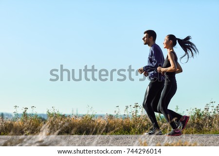 Young woman and man running along rural road. Athletic girl and guy in sportswear on morning jog outdoors. Photo with copy space on blue sky background. Full-length portrait of two sports people