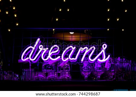 Glowing Purple Neon Inscription DREAMS and glasses on blurred lights background. Dark tones vintage image.