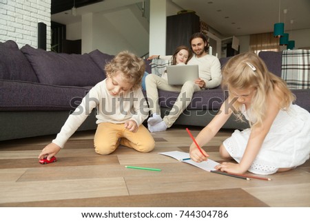 Kids siblings playing on floor while parents using laptop at home, preschool boy holding toy car, little girl drawing with colored pencils in album, family spending time together in living room 