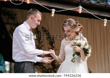 Beautiful and romantic wedding ceremony. Live white flowers, light bulbs, steps. Couple on the background of an arch. Exchange of rings. The bride is putting the ring on the groom's finger.