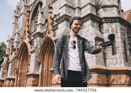 Cheerful cool and nice traveller is taking a selfie. Besides him there is a big great church building. Young man is smiling. He looks happy.
