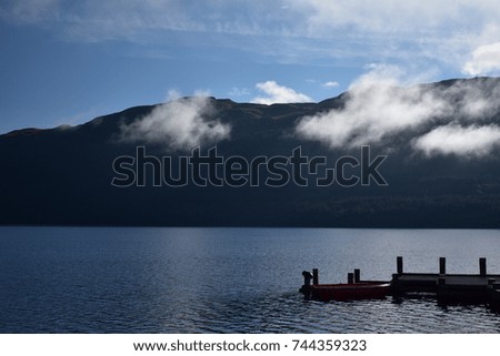 An image taken of a red boat tied at a dock with a glassy water and mountains in the background taken at Loch Lomond and the Trossachs National Park in Scotland in the United Kingdom.