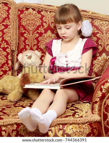 Beautiful little blonde girl with short bangs and pigtails on her head in a good mood.She plays with a teddy bear.She sits on the couch and reads the book.