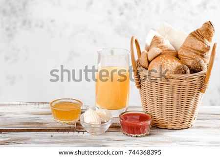 Baking breakfast croissants and bun in basket with orange juice, honey, butter, raspberry jam on white wood table