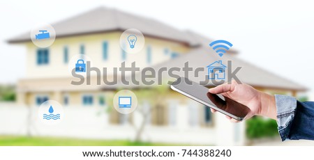 Hand using smart phone as smart home control application over blurred house background, banner, smart home concept Royalty-Free Stock Photo #744388240