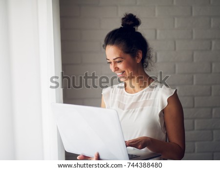40 year old smiling businesswoman holding a laptop while standing near the window.