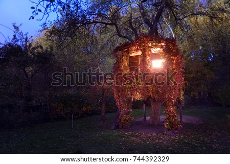 A house on a tree luminous from the inside and overgrown with red vine in an autumn garden