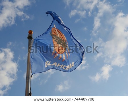 The flag of Oklahoma State waving in the wind with clear blue skies and clouds in the background