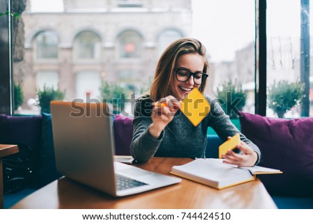 Positive hipster girl in stylish spectacles for better view using stickers for productive learning information from book.Smiling student sitting indoors at laptop computer preparing for upcoming exams Royalty-Free Stock Photo #744424510