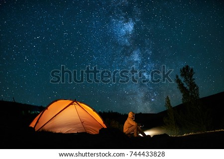 Man tourist near his camp tent at night under a sky full of stars. Orange illuminated tent.