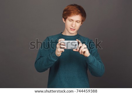 Close up of young good-looking redhead teenage boy with short hair in green sweater holding smartphone in hands, trying to make photo of friends in university. Black background