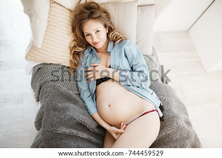 Portrait of attractive young joyful pregnant woman with blond curly hair in comfy home wear lying in her bed in morning, smiling happily in camera, touching stomach with hands.