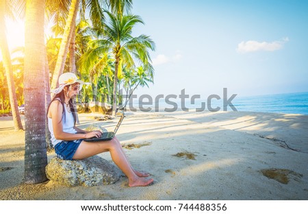 A woman using a laptop computer under coconut tree during holiday. Asia lady traveler have a relax time on the beach.
