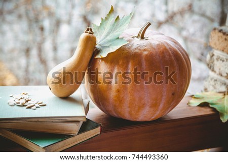 Beautiful romantic autumn still life. Two pumpkins of different shapes and a stack of books lie on the railing of the veranda. On top are seeds and a maple leaf
