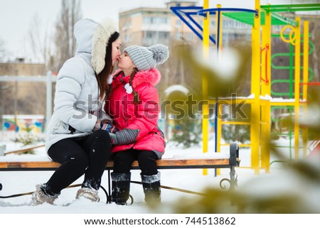 Two happy girls, mother and daughter sitting on a bench on a  playground in city park  at winter frosty day.
