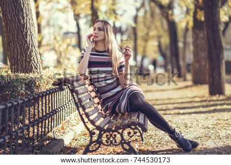 Young blonde woman sitting on a bench in the street of a park with autumn colors. Beautiful girl in urban background wearing striped dress. Female with straight hair holding an autumn leaf.