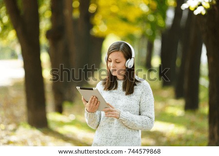 The beautiful happy cheerful brown-haired woman in white sweater with a tablet listening music in the white headphones in fall park on a warm day. Autumn in the city.