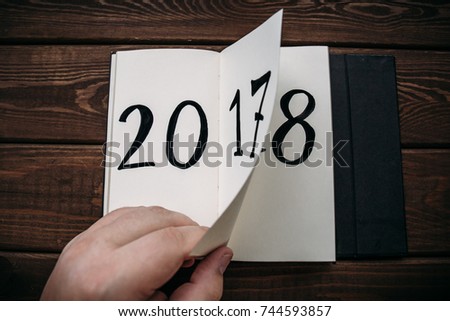 New Year 2018 is coming concept. Hand flips notepad sheet on wooden table. 2017 is turning, 2018 is opening. Top view,
