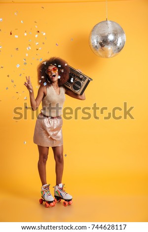 Full length portrait of overjoyed afro american disco woman in sunglasses showing peace gesture, standing on roller skates, holding boombox, looking at camera, isolated on yellow background Royalty-Free Stock Photo #744628117