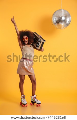 Full length portrait of happy afro american disco woman in sunglasses pointing with finger upward, standing on roller skates, holding boombox, looking at camera, isolated on yellow background