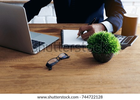 Portrait of handsome successful businessman working with laptop sitting at big desk in modern office, writing something in planner