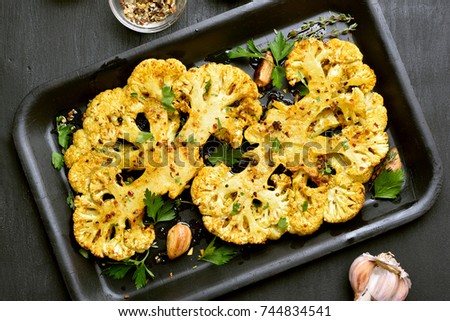 Baked cauliflower steaks with herbs and spices on baking sheet over black stone background. Royalty-Free Stock Photo #744834541