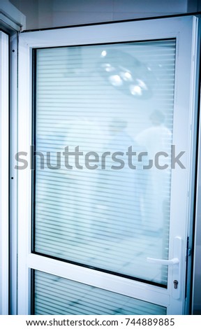 white door in operation room with reflection of doctors and patient. doctors stand around table with patient and big round lamp above him in reflection in door of operation room