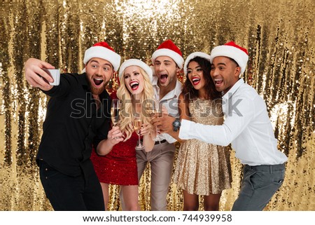 Portrait of a cheerful happy multiracial group of friends celebrating New Year together while drinking champagne and taking a selfie isolated over golden shiny background