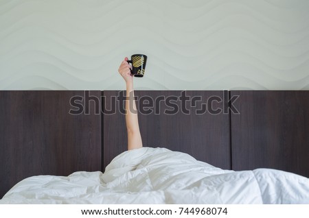 Drinking coffee early tomorrow. Woman in bed, under duvet, with arm raised up holding coffee cup. Royalty-Free Stock Photo #744968074