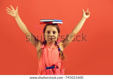 Back to school concept. School girl with funny face holds hands up, isolated on red background. Girl holds red and blue books on head. Pupil in pink dress and bag wears pencils and markers in hair