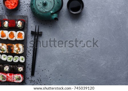 Top view on sushi set at gray background with scattered black sesame. Slate platter with rolls, ginger and soy sauce, chopsicks on rustic surface. Japanese restaurant food and delivery, copy space