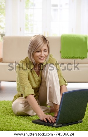 Smiling woman sitting on living room floor in front of sofa, working on laptop.?