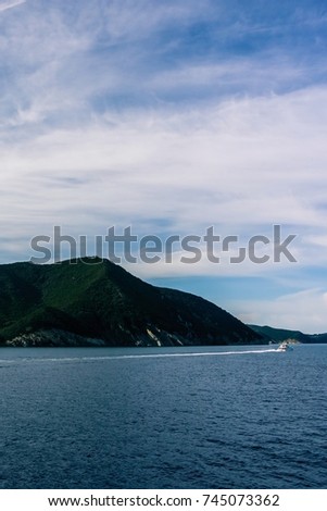Vertical photo of a coastline of island and boat in Mediterranean Sea. The coast is with several rocks of mountains in Tuscany Italy. The rocks are on Elba island. Sky is blue with few clouds.