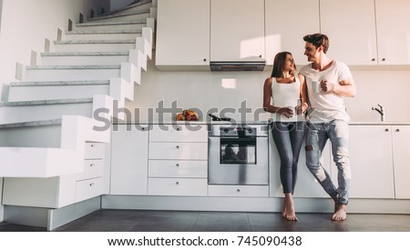 Full-length image of romantic couple at home. Attractive young woman and handsome man are enjoying spending time together while standing on light modern kitchen with cup of coffee in hands.