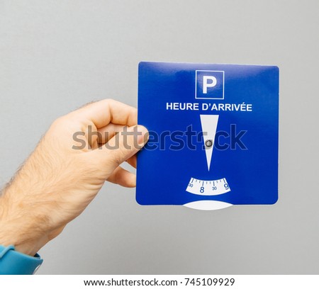 Man holding disk parking with french text Heure d'arrivee  translated as Arrival time from French language - gray background  