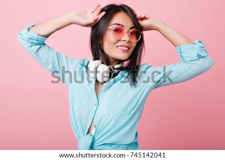 Romantic hispanic woman with straight hair standing with hands up and lovely smile. Indoor photo of magnificent asian girl in headphones and sunglasses stretching on pink background.