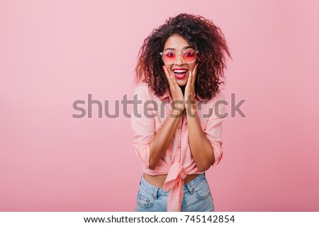 Terrific african model expressing surprised emotions while posing on indoor photoshoot. Stylish curly woman in sunglasses having fun in pink studio. Royalty-Free Stock Photo #745142854