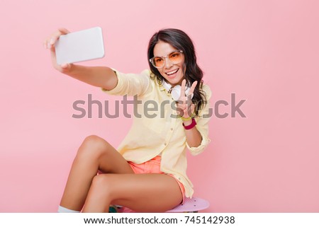 Good-looking european woman with bronze tan making selfie on smartphone. Glad lovable girl in yellow jacket taking picture of herself with peace sign.